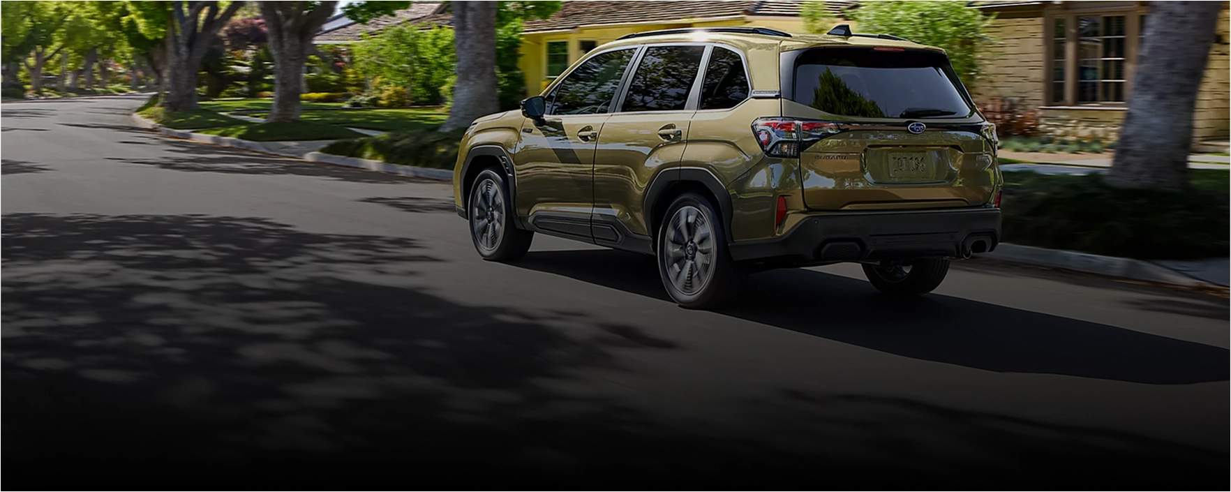 Green 2026 Subaru Forester Hybrid driving on a tree-lined residential street, rear three-quarter view