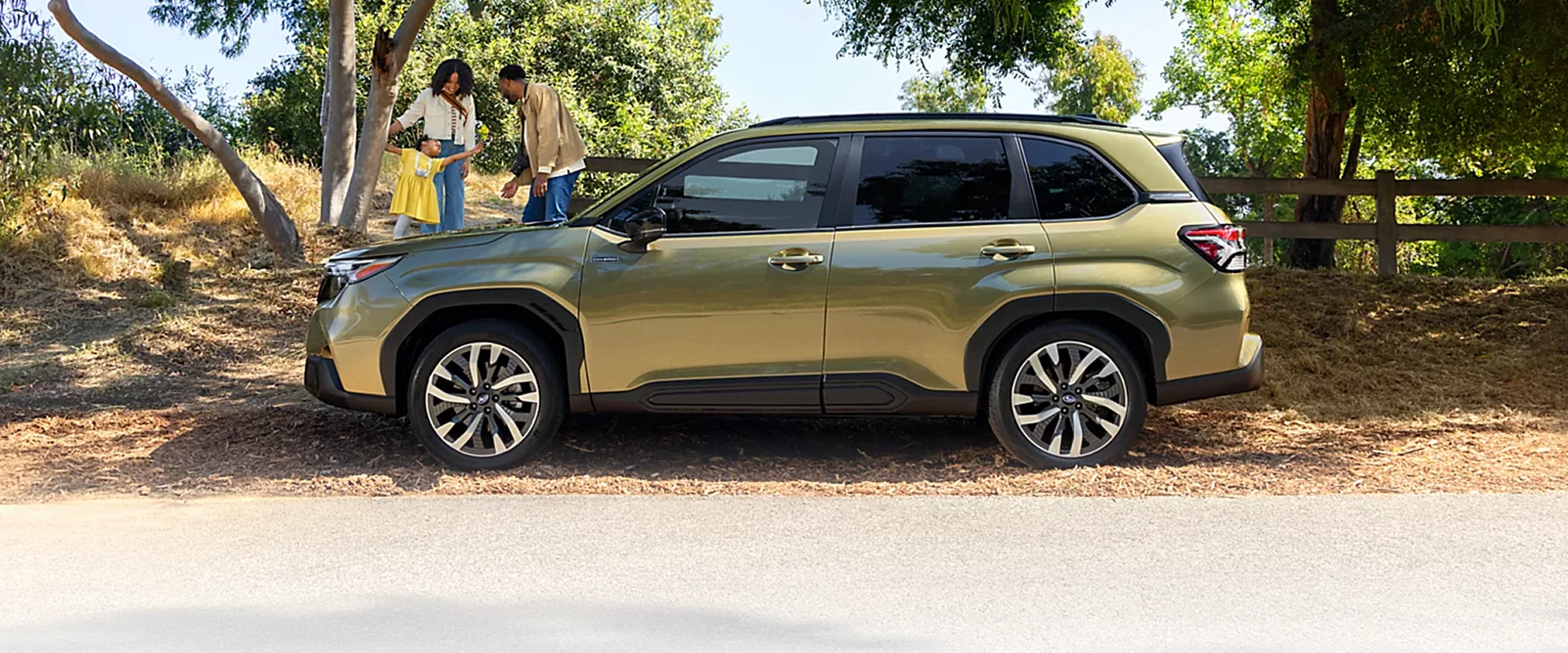Green 2026 Subaru Forester Hybrid parked on a tree-lined dirt path, side profile view, with two people standing beside it