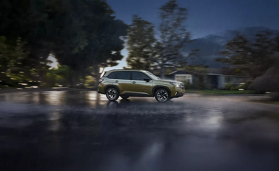 Green 2026 Subaru Forester Hybrid driving on a wet tree-lined road at dusk, side profile view