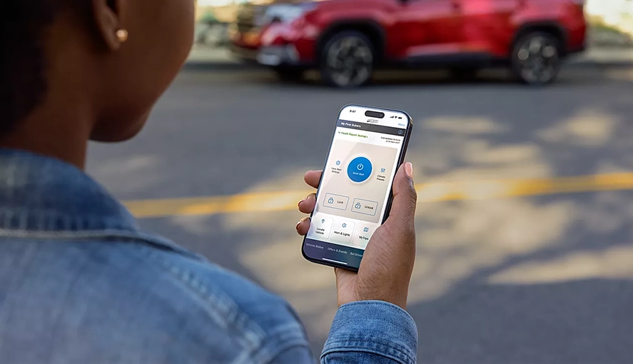 Person holding a smartphone showing the MySubaru Connected Services app, with a red 2026 Forester Hybrid in the background