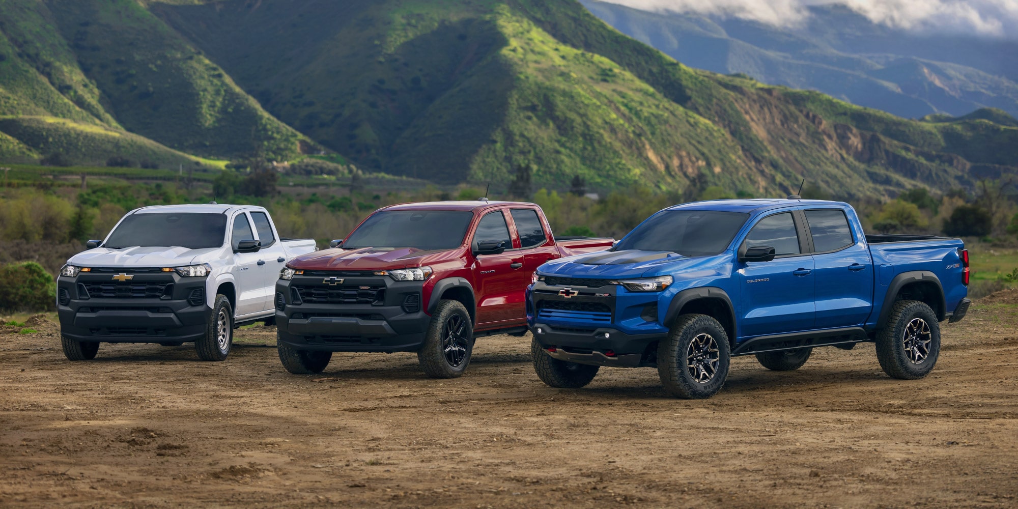 A trio of white, red, and blue Chevrolet Colorados parked on a dirt road near mountains A trio of white, red, and blue Chevrolet Colorados parked on a dirt road near mountains