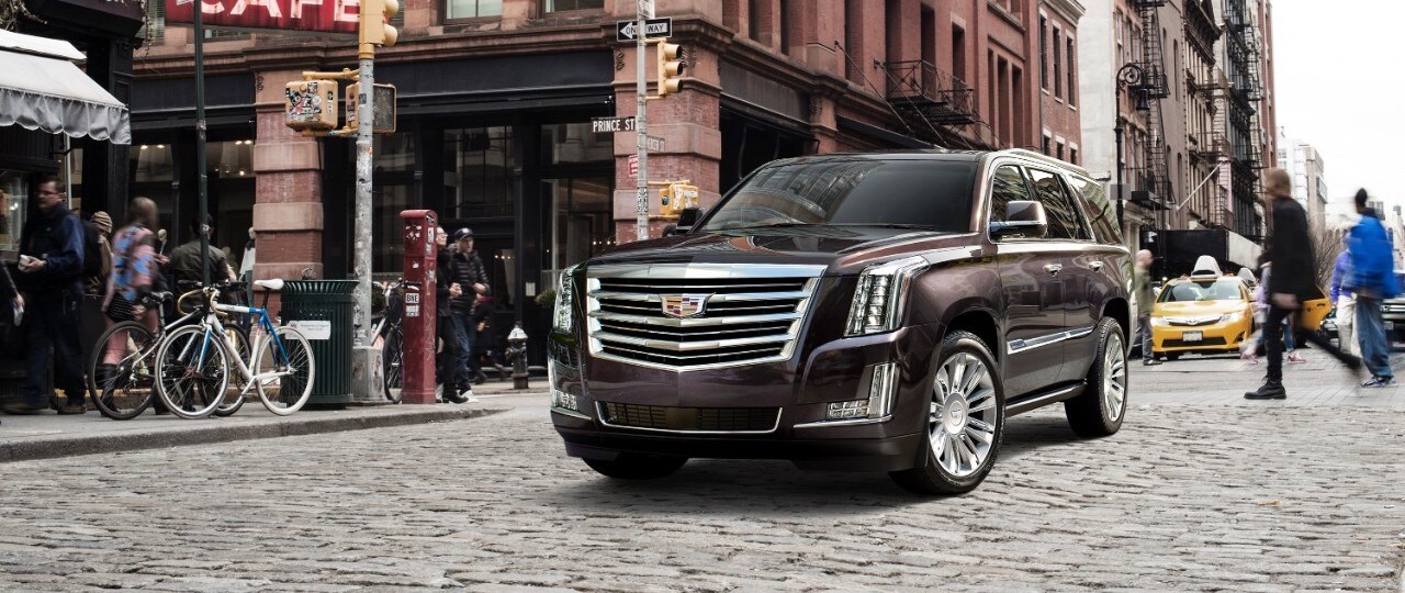 Cadillac Laval - VUS Cadillac d'occasion - Escalade Platinum 2017 - 0,4.jpg Cadillac Escalade Platinum 2017 garé dans une rue de centre-ville avec des piétons traversant la rue