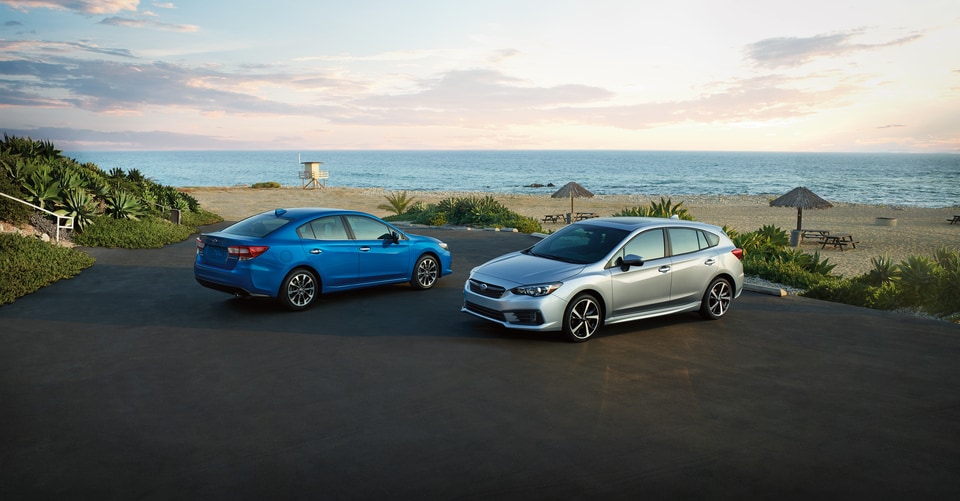 2021-Subaru-Impreza-Lineup-01.jpeg a blue Subaru Impreza sedan and silver Subaru Impreza hatchback parked next to a beach