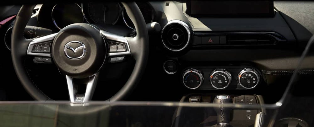 2023 Mazda MX-5 Miata Interior> </p><h2>2023 Mazda MX-5 Miata Interior</h2><p>The entry Sport trim's snug two seater cabin offers plentiful standard comfort features including, air conditioning, power windows with driver's and passenger's one-touch-down features and power door locks with a speed sensor.</p><p></p><p>Convenience features abound such as the side window demisters, dual vanity mirrors, overhead courtesy light, 12-volt power outlets plus top down travel is enhanced by the standard wind blocking mesh aero board.</p><p></p><p>The Sport trim has bucket seats with integrated headrests and a reclining and forward/back slide feature upholstered in black cloth, with the driver enjoying a tilt adjustment seat. The sporty three-spoke steering wheel is wrapped in leather along with the shift knob and parking brake handle.</p><p></p><p>Further key cabin finishes are padded armrests, black upper door trim, and carpet floor mats.  The locking rear center console, two removable cup holders, plus driver's and passenger's back panel-covered storage, offer plentiful cabin storage areas. </p><h2>Mazda MX-5 Miata Performance</h2><p>The <a href=
