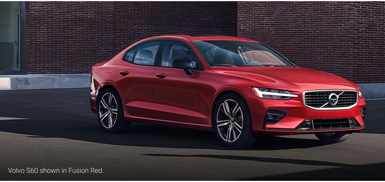 Front three-quarter view of a 2026 Volvo S60 in Fusion Red Metallic parked in front of a modern red brick building.