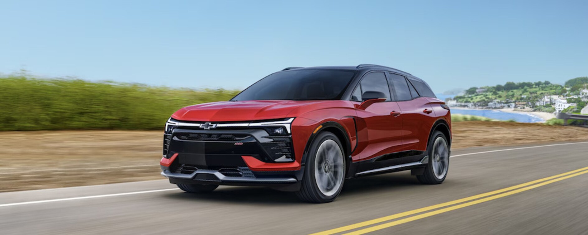 A red 2026 Chevrolet Blazer EV SUV driving down the road with a seaside town in the distance
