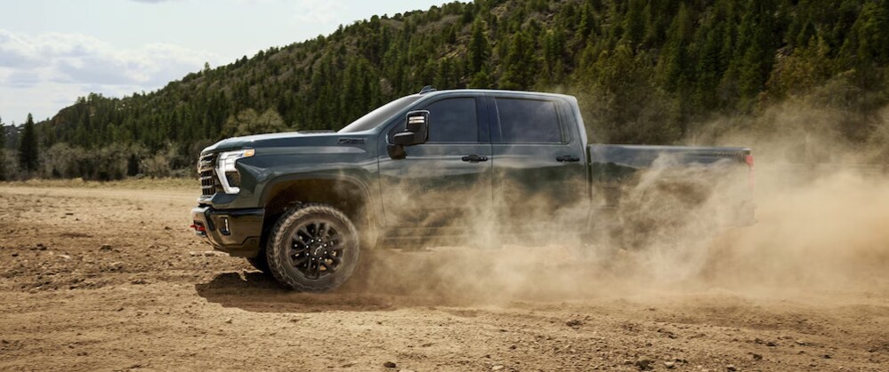 A dark-colored 2026 Chevrolet Silverado HD pickup driving on bare dirt, with a cloud of dust swirling around the truck