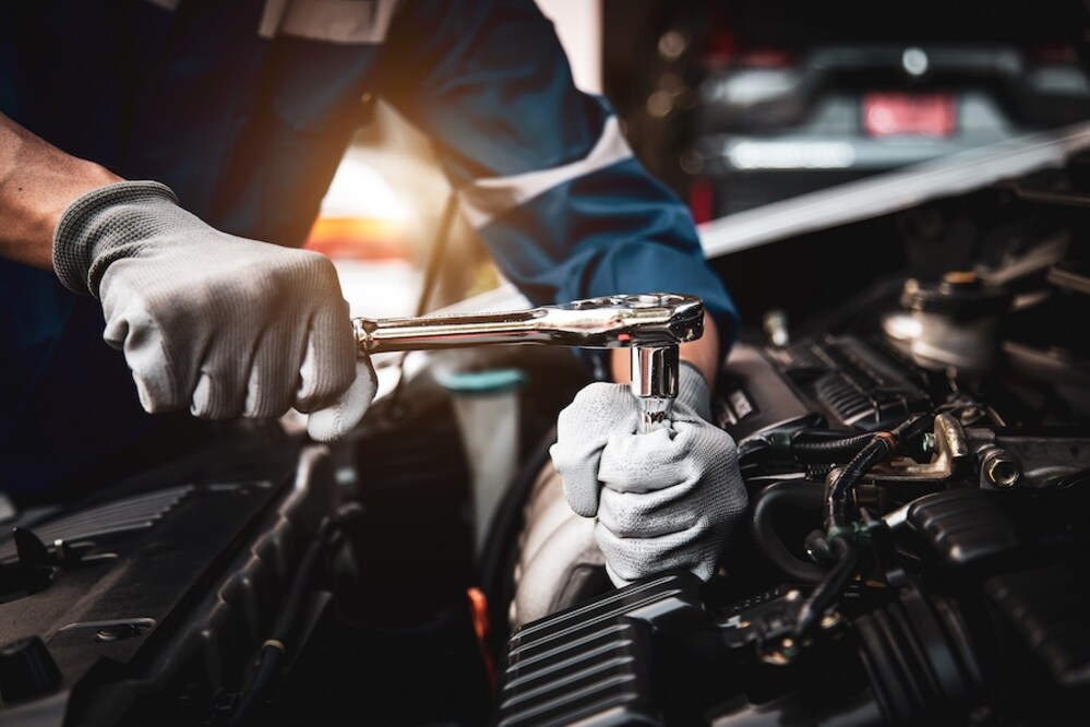 A person in gray gloves and blue coveralls working on a car's engine with a wrench