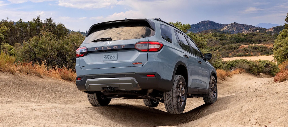 A light blue 2026 Honda Pilot SUV driving over uneven terrain in desert scrub, with mountains in the background