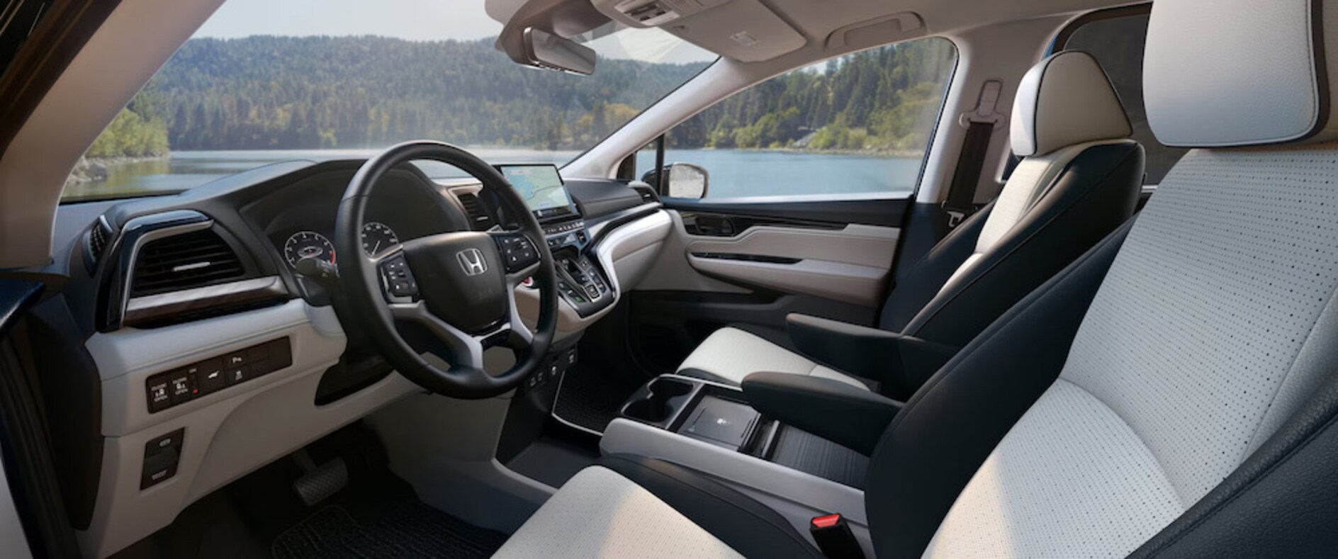 The front seats and dash of the 2026 Honda Odyssey van, featuring grey and white upholstery and a touchscreen display, with a tree-lined lake visible through the windows
