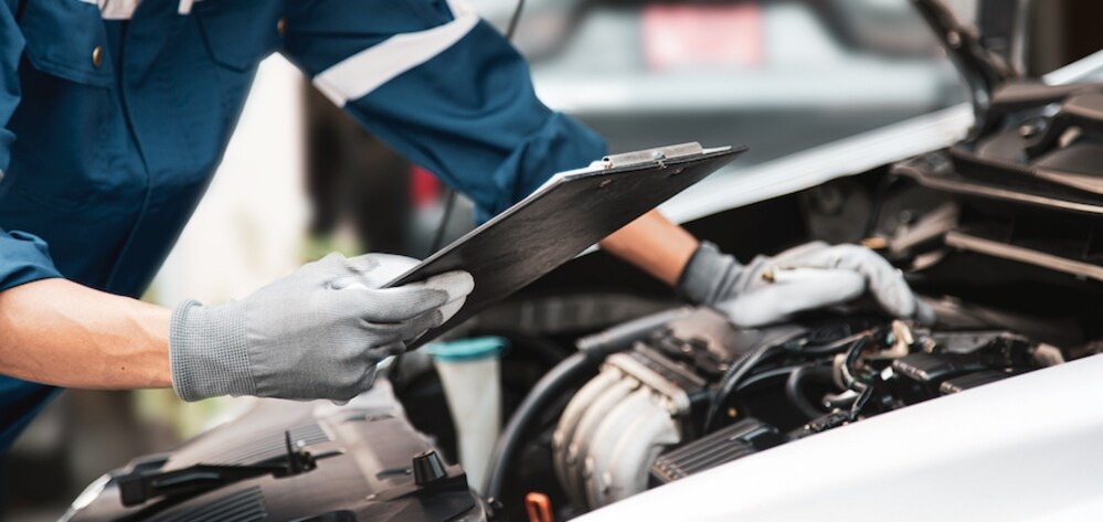 The torso of a white person in blue coveralls and gloves holding a clipboard and looking at the engine of a vehicle