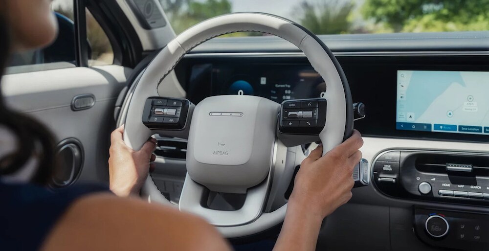 The shoulder and hand of a dark-haired woman sitting in the driver's seat of a 2026 Hyundai Palisade hybrid SUV, with a white steering wheel and navigation visible on the display The shoulder and hand of a dark-haired woman sitting in the driver's seat of a 2026 Hyundai Palisade hybrid SUV, with a white steering wheel and navigation visible on the display