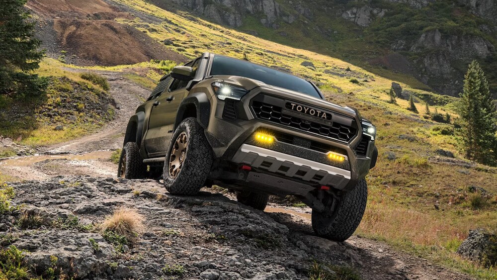 An olive green 2026 Toyota Tacoma pickup truck driving up a rocky outcropping An olive green 2026 Toyota Tacoma pickup truck driving up a rocky outcropping