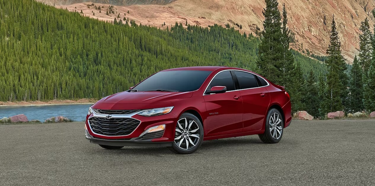 2023-chevy-malibu-3.PNG A parked red 2023 Chevy Malibu parked in front of a mountain lake in profile view