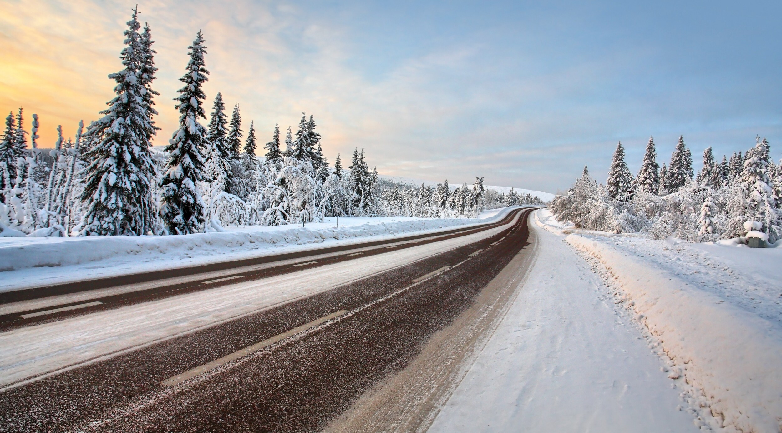 AdobeStock_49126200.jpeg A snow packed road at sunset surrounded by forests .