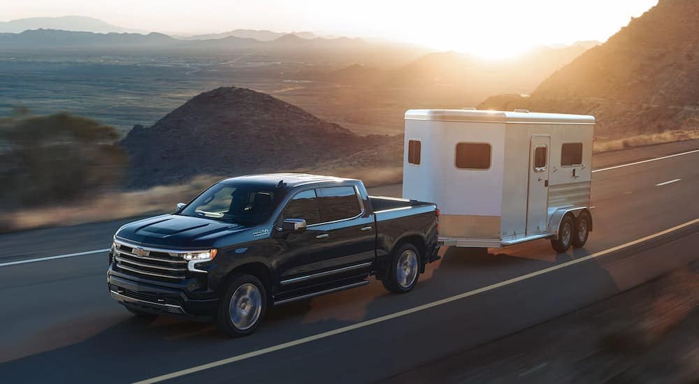 A black 2023 Chevy Silverado 1500 High Country is shown towing a white trailer on an open road.