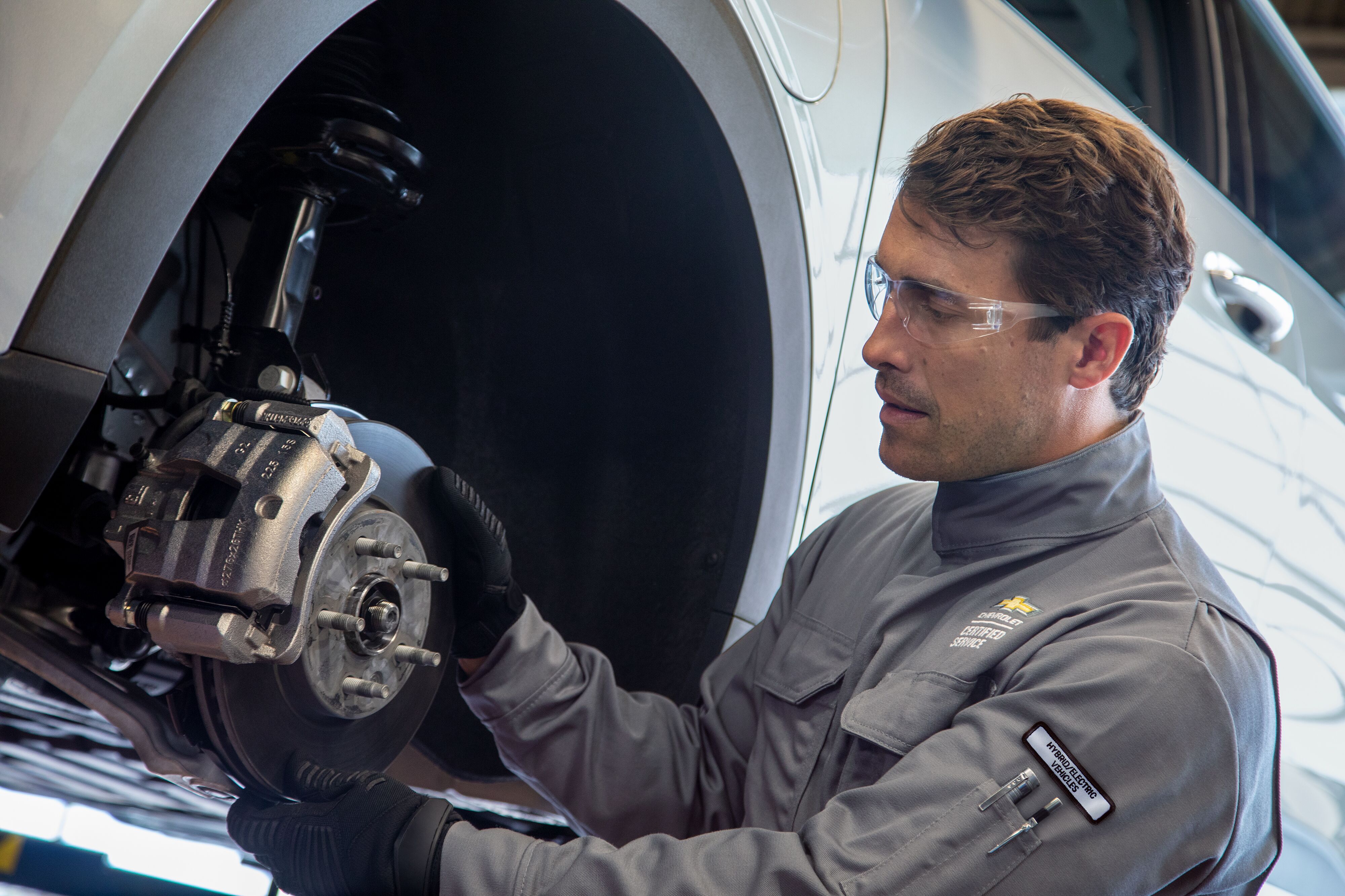 Chevy Maintenance Certified service mechanic wearing safety glasses inspecting brake rotor and caliper on an elevated hybrid electric vehicle.