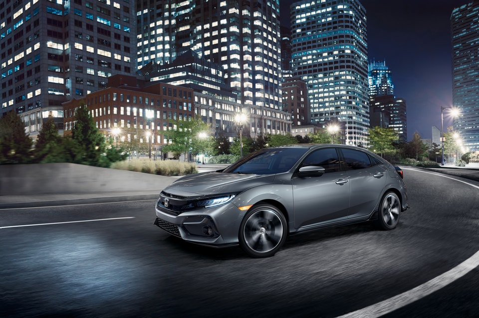 2022-Honda-Civic-SportTouring-Hatchback-L01.png dark gray Honda Civic Hatchback driving through a well-lit downtown area at night