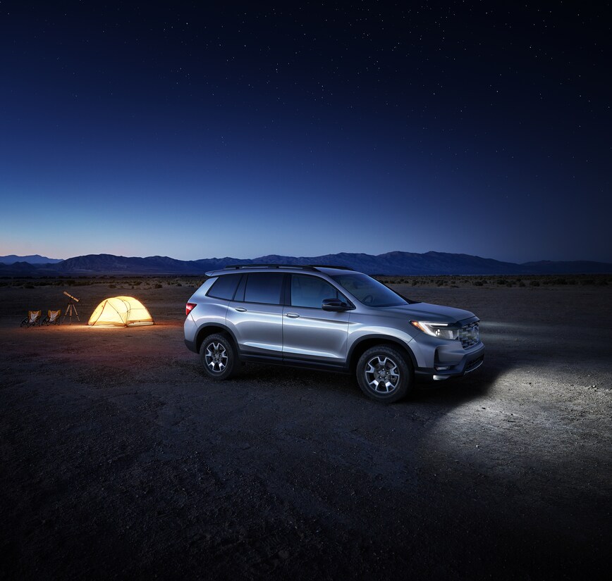 Honda-Passport-TrailSport-SUV-L13.png silver Honda Passport SUV parked in the desert with its headlights on and a lit tent nearby