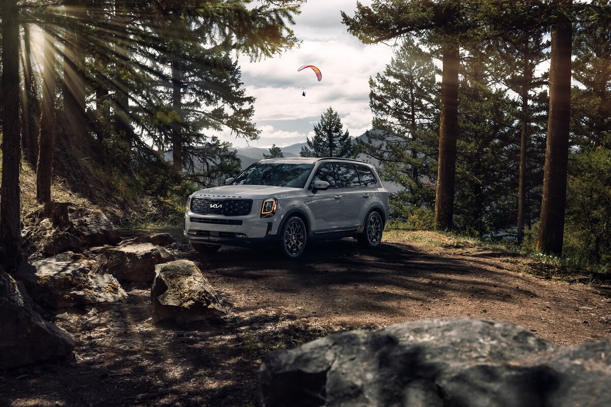 2022-KIA-Telluride-SX-SUV-L01.png silver Kia Telluride SUV parked in a forest with a parasailer beyond