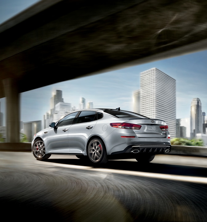 2019-KIA-Optima-SX-Sedan.png light silver Kia Optima car driving around a tight corner underneath a freeway overpass