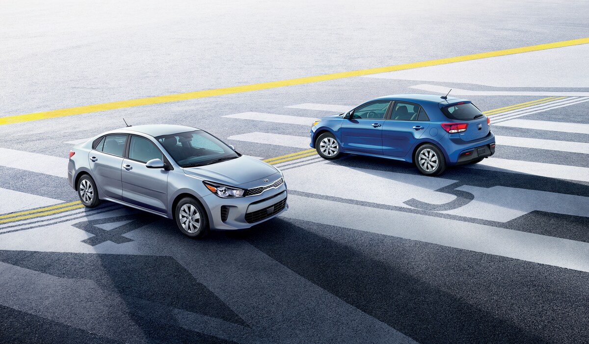 2020-KIA-Rio-S-Hatchback-L02.png silver and blue Kia Rio hatchback cars parked next to each other on a tarmac with the drivers facing each other out the side windows