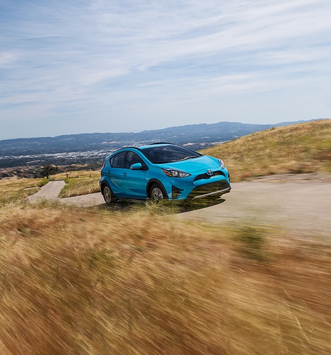 2019-Toyota-Prius-LE-Hatchback.png light blue Toyota Prius hatchback driving by a dry, grassy field