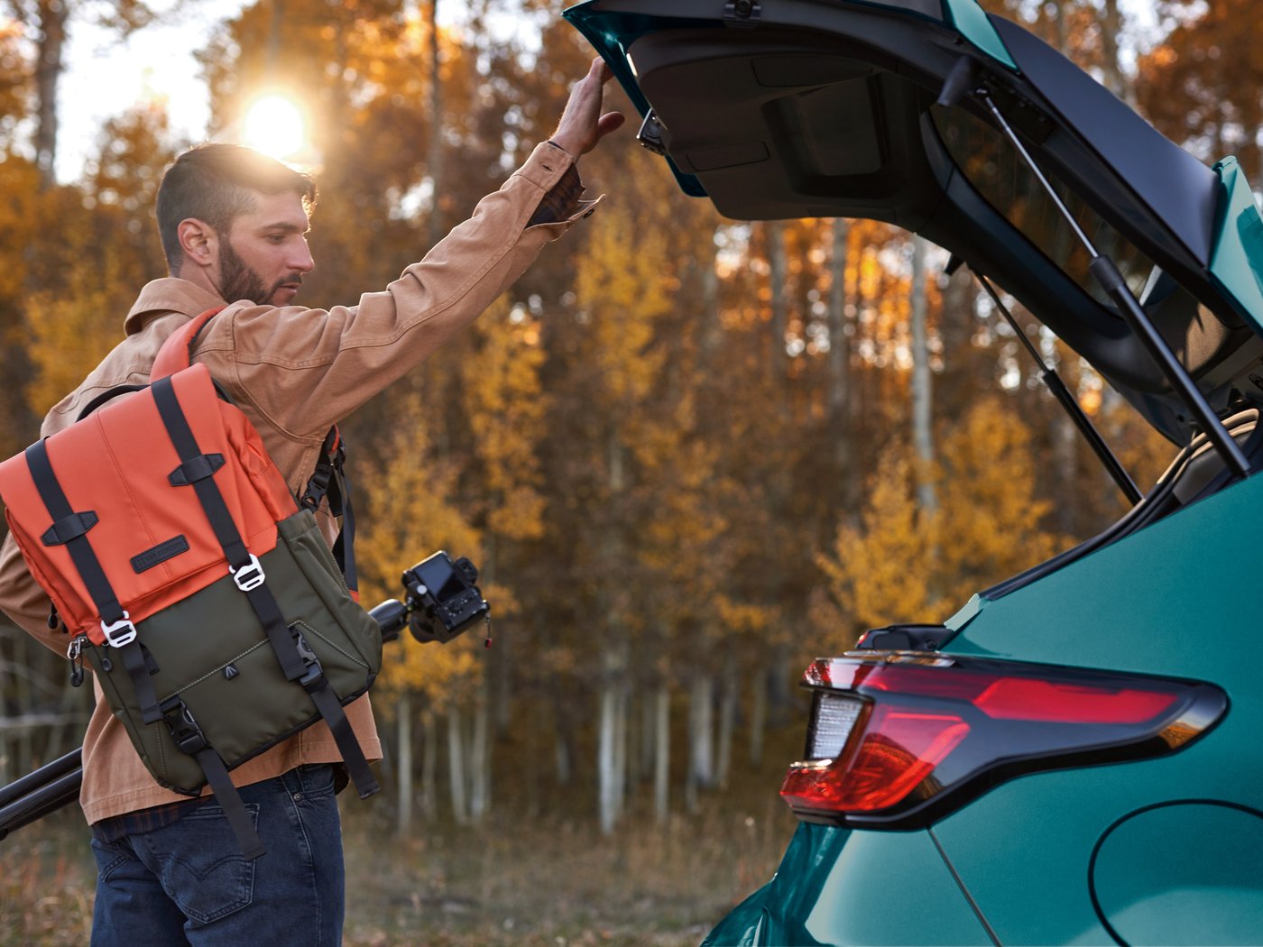 A man in a backpack lifts the liftgate of a Tidepool Teal Pearl Crosstrek among aspens in fall color