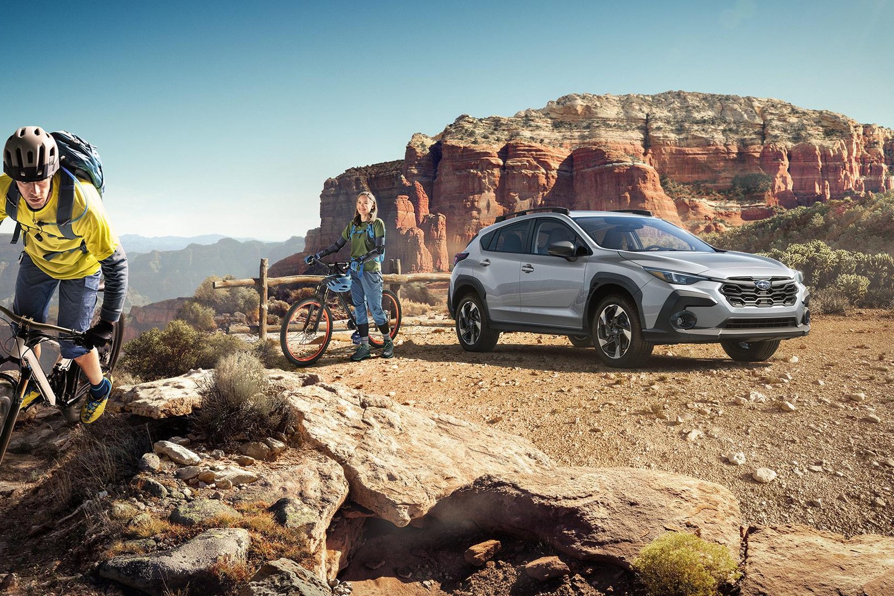 An Ice Silver Metallic 2027 Subaru Crosstrek parked on a rocky overlook in red-rock country, with two mountain bikers beside it