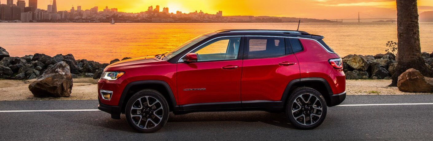2019 Jeep Compass exterior side shot with red paint color parked outside near a lake and city and a beautiful sunset
