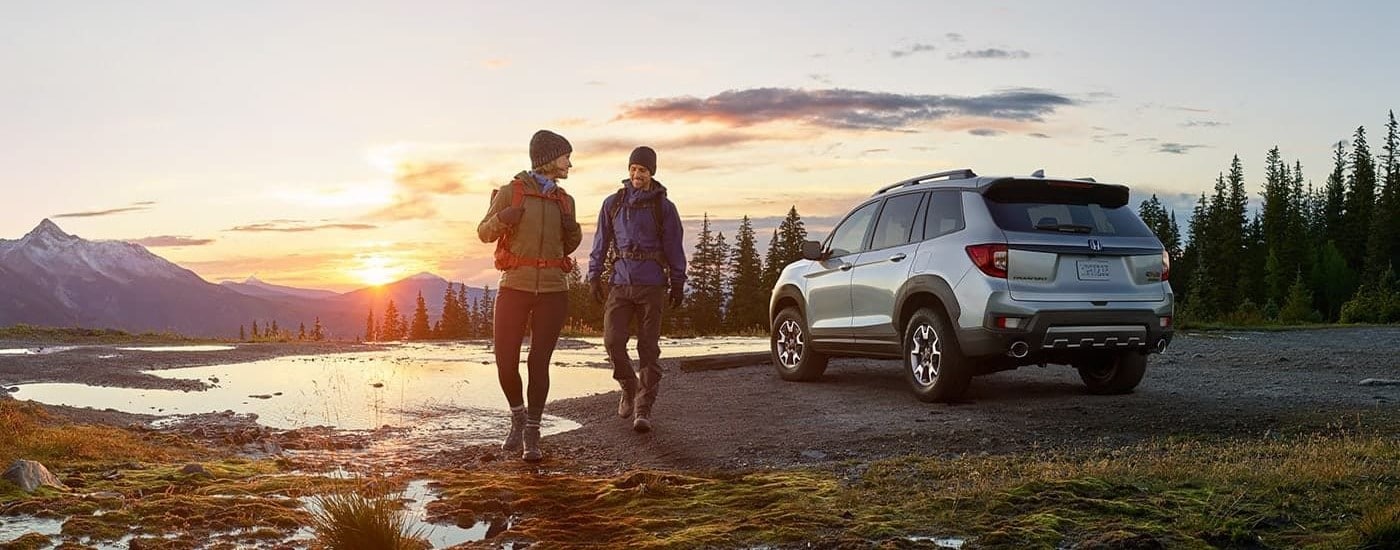 Honda SUVs for Sale - 2023 Honda Passport TrailSport - Sunrise.jpg A silver 2023 Honda Passport TrailSport is shown at sunrise near hikers.