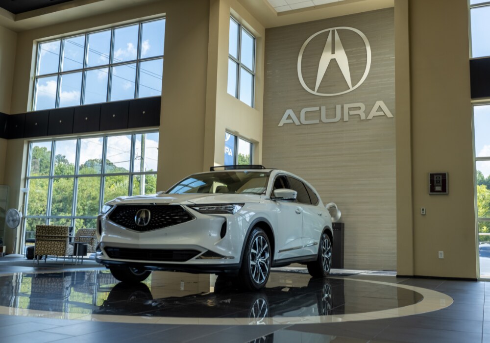 acura-dealership-serving-greensboro.png A white Acura SUV parked inside the Flow Acura dealership in Winston-Salem, behind the car, there is a large Acura logo affixed to the wall