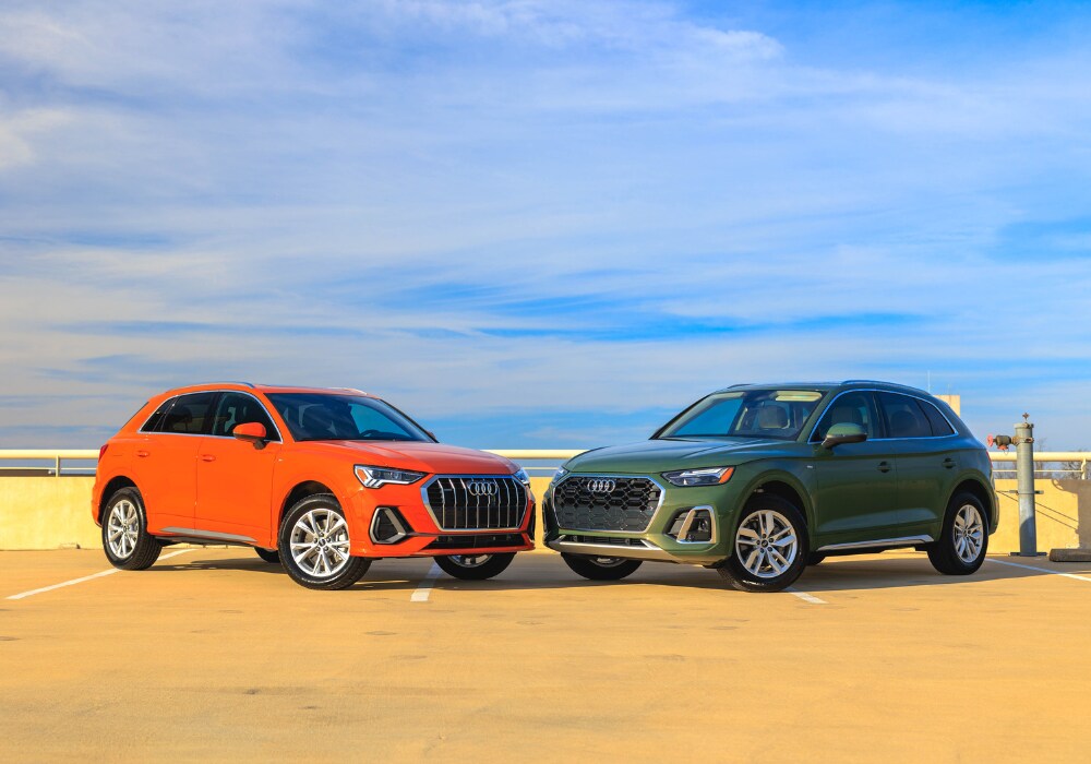 audi-suv-charlottesville.png Two 2024 Audi SUVs parked on a paved lot in front of a blue sky. The Audi SUV on the left is orange and the Audi SUV on the right is green