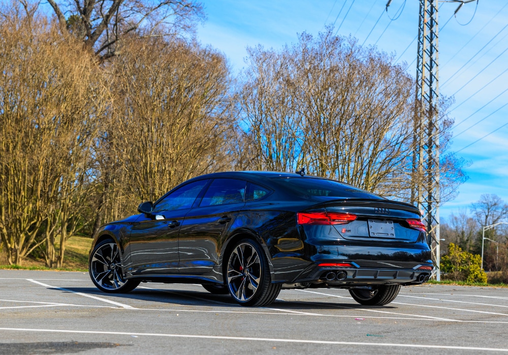 2024-audi-s5-greensboro.png A black 2024 Audi S5 Sportback parked in an empty parking lot with a clear blue sky and transmission towers in the background. The car is facing away from the camera, showing its rear end and red brake lights.
