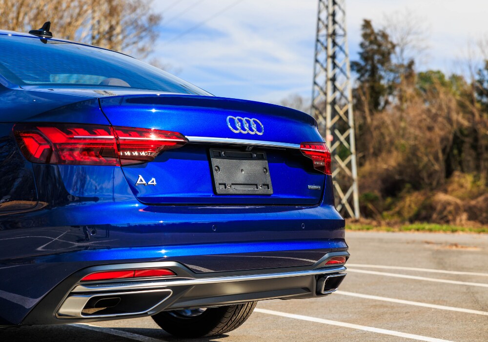 audi-a4-quattro.png A blue Audi A4 sedan parked in a parking lot with a clear sky and trees in the background. The car's rear end is visible, showing the Audi logo, taillights, and a chrome accent on the bumper