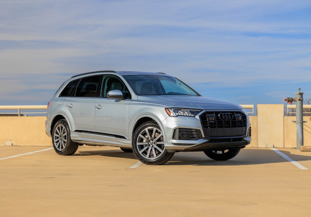 audi-q7-for-sale.png Silver 2024 Audi Q7 SUV parked on top of a parking garage with a blue sky in the background