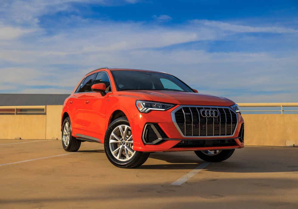 2024-audi-q3.png A bright orange 2024 Audi Q3 SUV parked on a rooftop in Greensboro with a clear blue sky in the background.