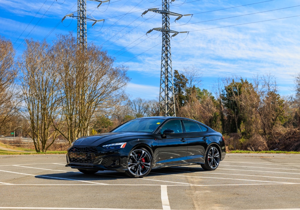 ausi-s5-for-sale.png A black 2024 Audi S5 Sportback parked in an empty parking lot in Greensboro with a clear blue sky and transmission towers in the background.