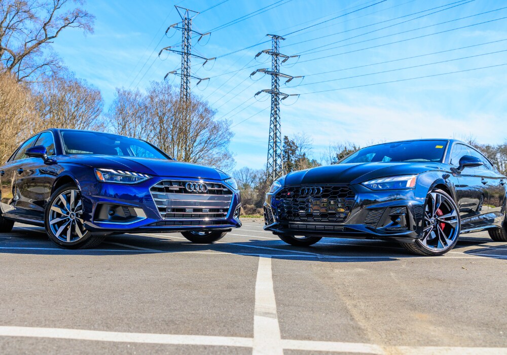 audi-sedans-for-sale.png Two Audi cars parked side-by-side in a parking lot. The car on the left is blue and appears to be a 2024 Audi A5. The car on the right is black and appears to be a 2024 Audi S5 Sportback. Both cars are facing the camera with a clear blue sky and transmission towers in the background