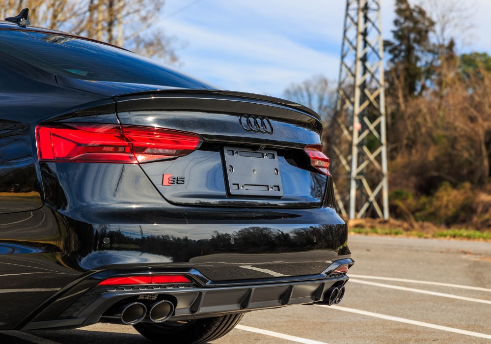 audi-s5-sportback.png A close-up of the rear of a black 2024 Audi S5 Sportback. The car has a spoiler, red taillights, and quad exhaust tips.