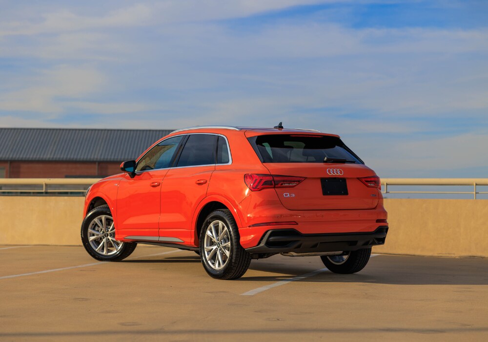 audi-q3-for-sale.png A bright orange 2024 Audi Q3 SUV parked on a rooftop with a clear blue sky in the background. The photo shows the rear of the vehicle with the Audi logo visible