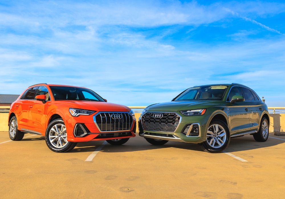 new-audi-suvs-for-sale.png A photo of two Audi SUVs parked side-by-side. The car on the left is orange and is a 2024 Audi Q3. The car on the right is green and is a 2024 Audi Q5. Both SUVs are parked in a parking lot with a clear blue sky in the background.