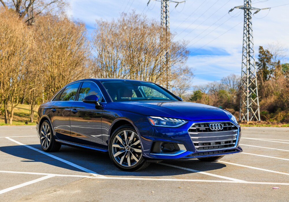 audi-a4-for-sale-greensboro.png A blue 2024 Audi A4 sedan parked in a parking lot with bare trees and power lines in the background. The car's front is visible, showing the Audi logo, headlights, and chrome grille