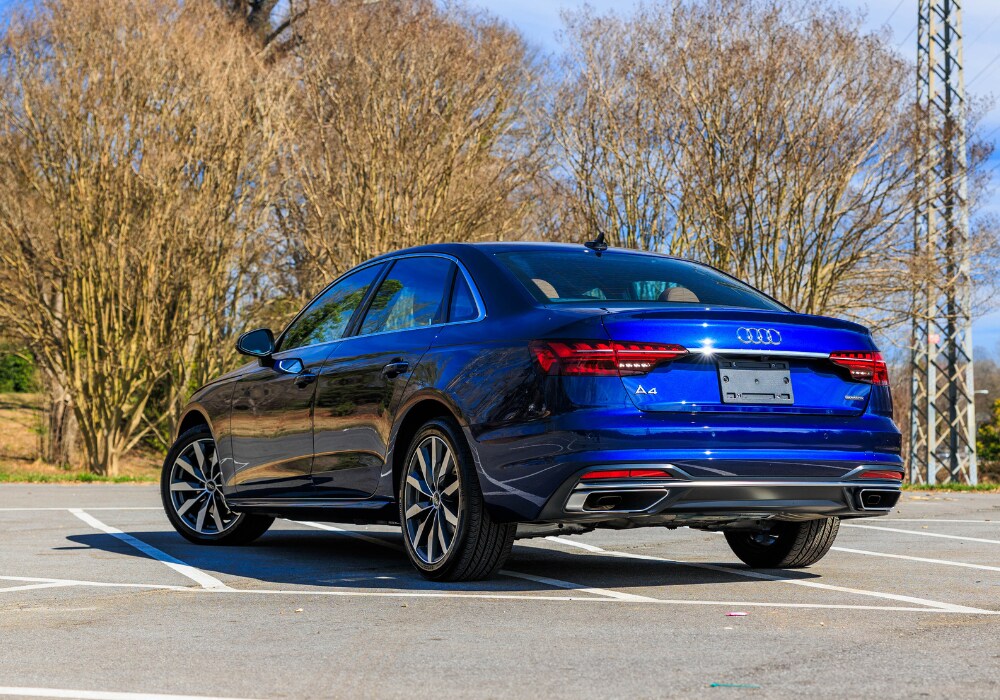 2024-audi-a4.png A blue 2024 Audi A4 sedan parked in a parking lot with bare trees and power lines in the background. The car's rear end is visible, showing the Audi logo, taillights, and a chrome accent on the bumper.