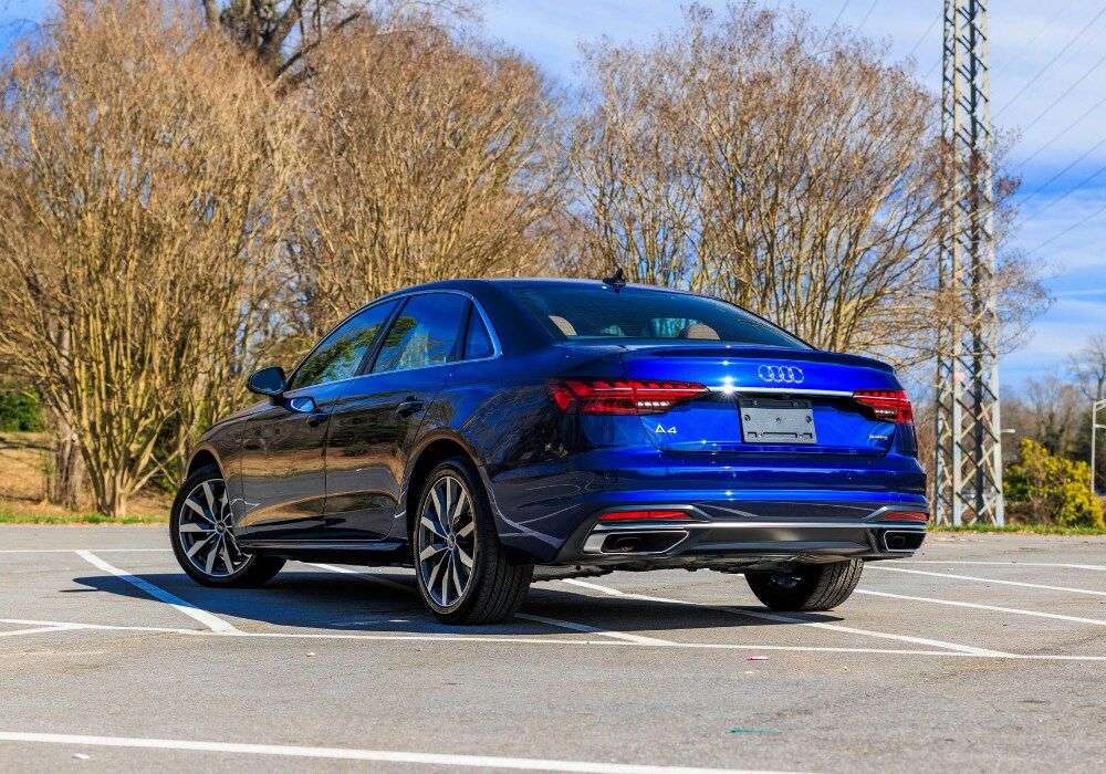 2024-audi-q4.png A blue 2024 Audi A4 sedan parked in an empty parking lot. The car is facing away from the camera, and its rear is visible. The background shows trees and a clear sky.