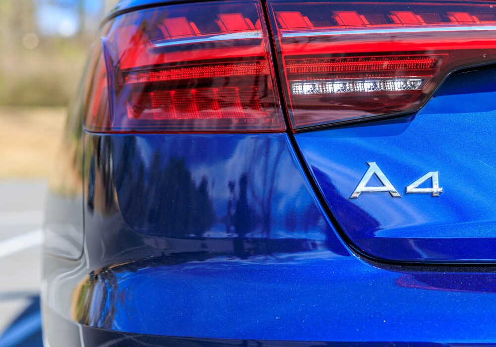 audi-a4-quattro.png A close-up of the rear of a blue 2024 Audi A4 sedan. The image shows the taillight with its red and white lights illuminated, as well as the Audi A4 badge.