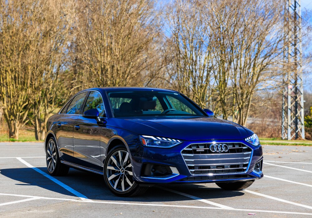 audi-a4-for-sale.png A blue 2024 Audi A4 sedan parked in a parking lot in Winston-Salem. The car is facing the camera, showing its front. There are trees and a clear sky in the background.