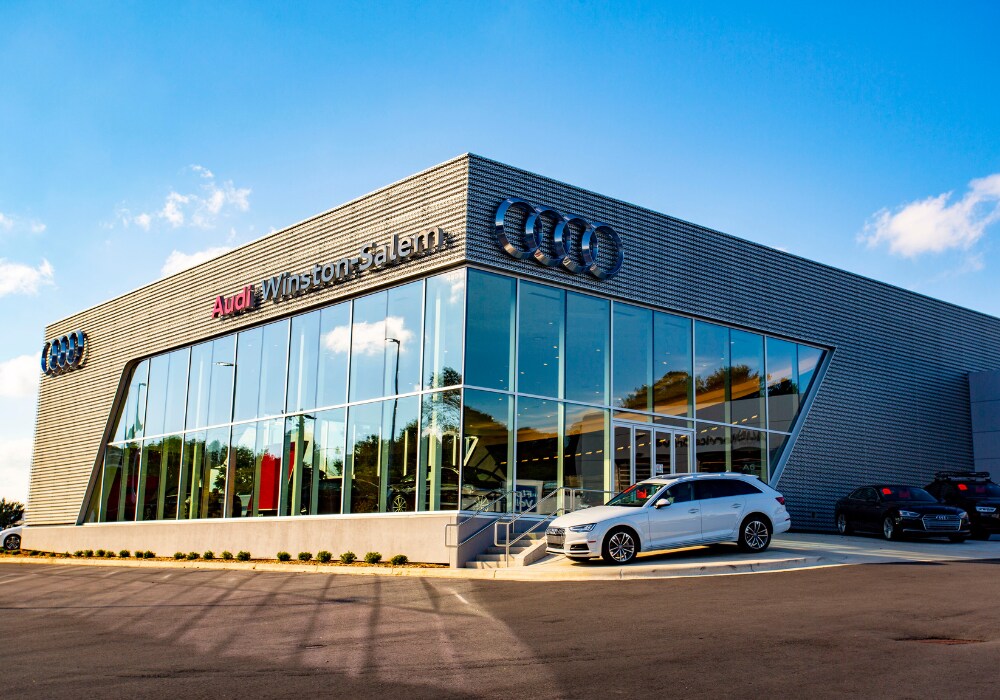used-audi-for-sale.png Audi Winston-Salem dealership storefront. The Audi logo is affixed to the building and there is a blue sky in the background. There is also a white Audi SUV for sale parked in front of the dealership