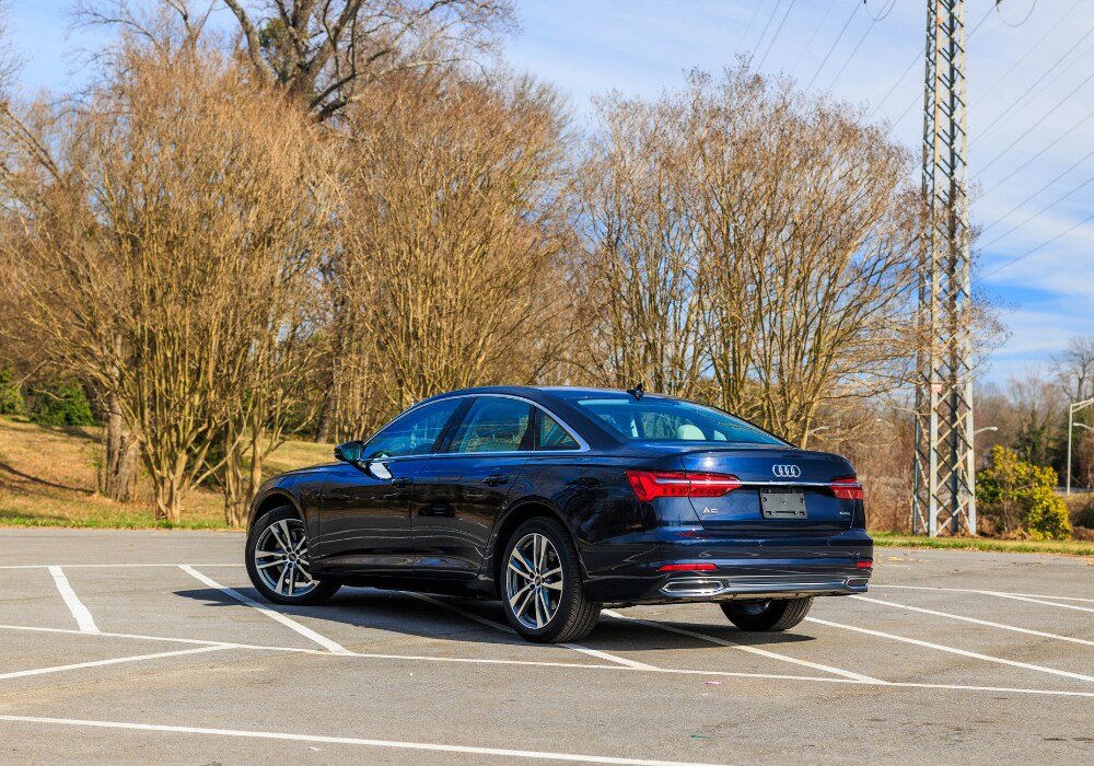 audi-a6-for-sale.png A blue 2024 Audi A6 sedan parked in a parking lot in Winston-Salem. The car is facing away from the camera, and its rear is visible. The background shows trees and a clear sky.