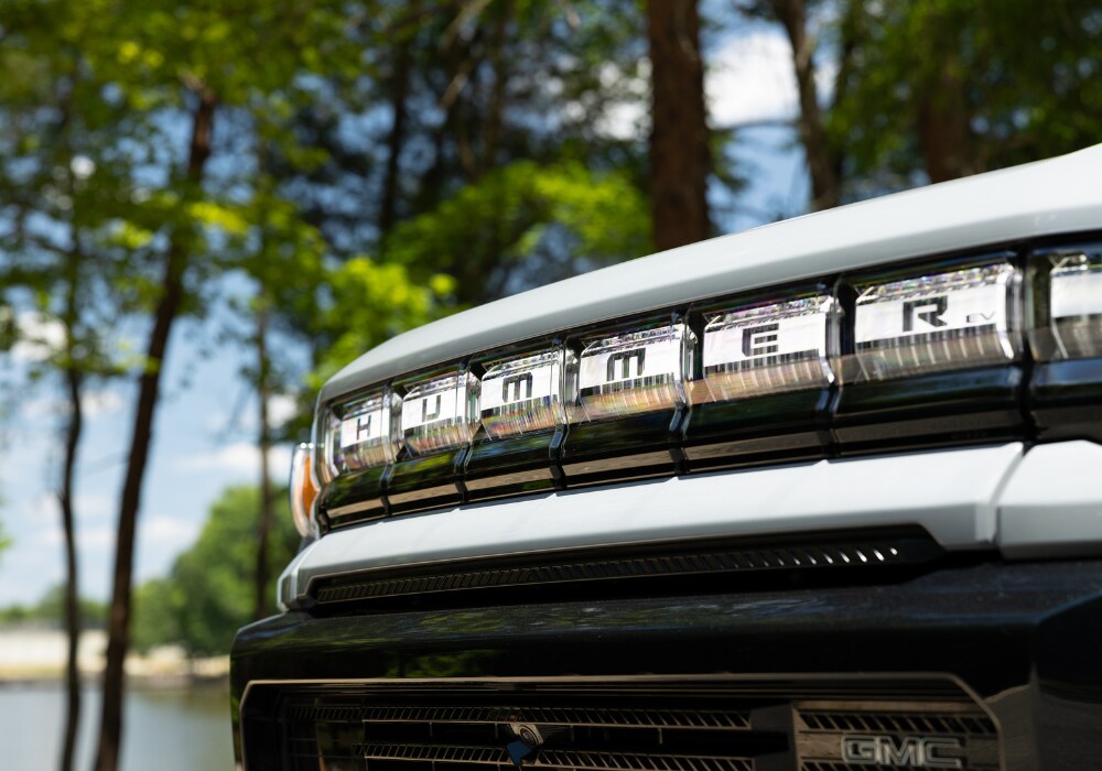 hummer-ev-suv.png Close up shot of the front grille of a 2024 Hummer EV SUV for sale in Winston-Salem. There are trees and a lake out of focus in the background